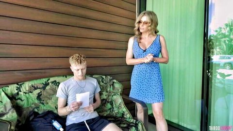 Cory Chase in sexy blue dress stands over young redhead guy on couch in taboo scene