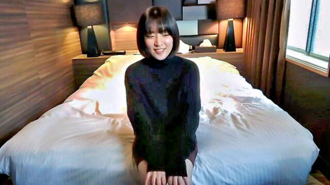 Cute Asian amateur in black turtleneck and skirt sitting on bed smiling