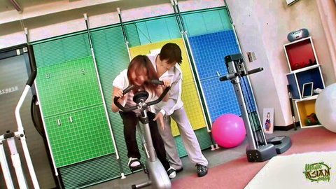 Chinese gym babe in tight pants pedals stationary bike while trainer grips her hips from behind