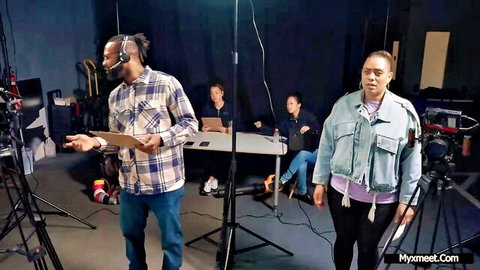Black host in plaid shirt gesturing during studio podcast interview with seated guests and standing woman