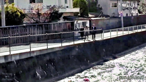 Two people walking along a fenced riverside path in urban Japan with apartments and trees