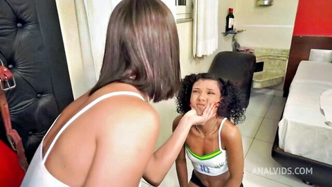 Brunette in white tank top leans over curly-haired Latina puckering lips teasing blowjob in bedroom