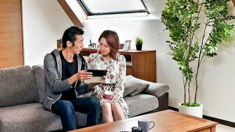 Young Japanese couple in casual clothes sit intimately on couch with man showing woman a tablet