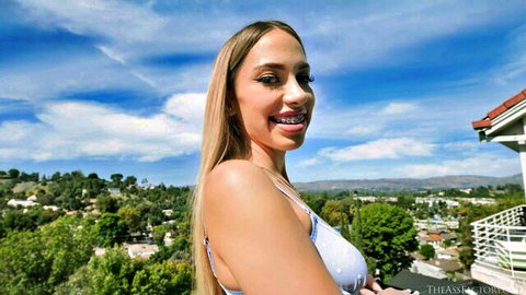 Khloe Kapri blonde smiling with braces on sunny balcony in white top