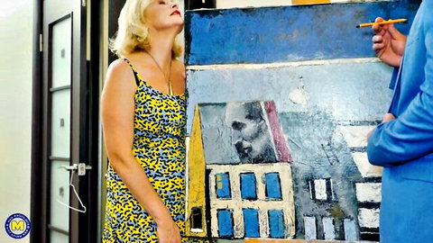 Blonde in tight yellow leopard dress with cleavage stands by painting with suited man holding pencil