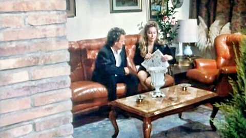 Moana Pozzi in black dress sitting with man on couch, 1992 adult scene