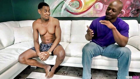 Two muscular black men sitting on couch, one shirtless in shorts gesturing casually in taboo scene