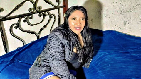 Timid Brazilian woman in black jacket and skirt sits seductively on blue bed
