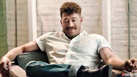 Curly-haired muscular man smiles relaxed in white shirt and jeans sitting in modern chair