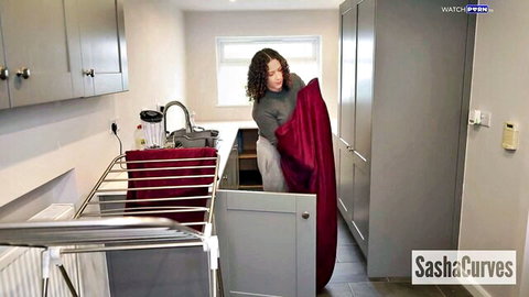 Curvy brunette Sasha Curves in grey sweater bending over holding red sheet in laundry kitchen