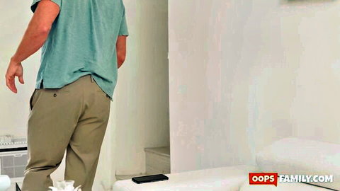 Clothed man in teal shirt and khaki pants walks towards white bed in bedroom