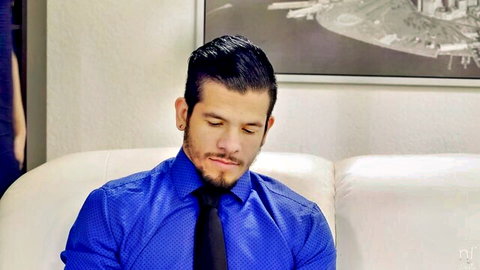 Handsome bearded man in blue polka-dot shirt and black tie sits pensively on white couch