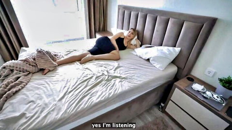 Blonde pregnant woman in tight black dress lying sideways on bed listening intently