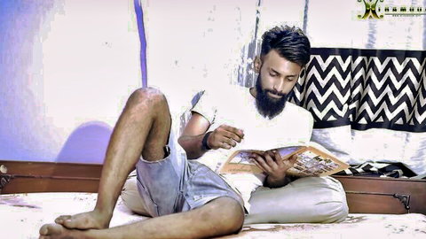 Bearded South Asian man lounging on bed reading book in tight shorts spreading legs