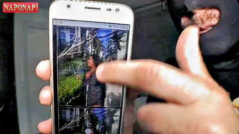 Man's hand holding smartphone screen showing brunette in dress amid jungle plants with SNAPNAP watermark