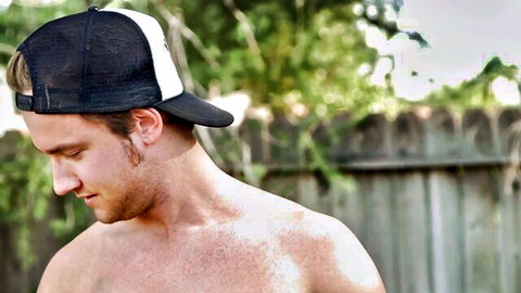 Sexy shirtless white hunk in trucker hat smirking seductively outdoors by fence