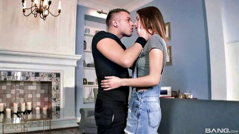 Tattooed guy passionately kisses sexy brunette in denim overalls by fireplace