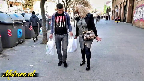 Mature blonde cougar in skirt stockings walks with young stud holding shopping bags on graffiti street