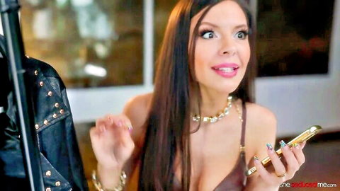 Darcie Dolce in low-cut top holding gold phone, smiling seductively