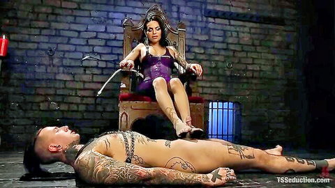 Tattooed brunette trans domme in purple latex throne holds crop over naked chained male slave on dungeon floor