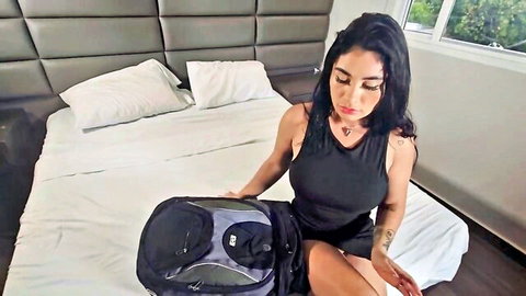 Tattooed Latina in black tank dress holds backpack on bed, teasing stepmom vibe