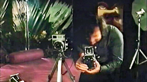 Vintage brunette in green outfit holds camera on tropical film set with tripods and palm plants