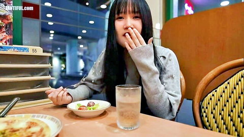 Young Asian girl in gray sweater covering mouth shyly at restaurant table with food and water
