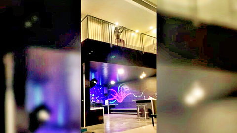 Thalia leans over balcony railing seductively with Felipe in purple-lit lounge