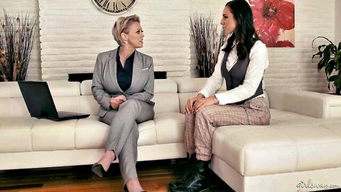 Blonde MILF in grey suit chats intimately with tanned brunette babe on white couch