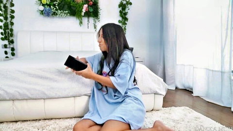 Petite Asian stepsister in loose blue dress kneeling on bed, teasing phone view