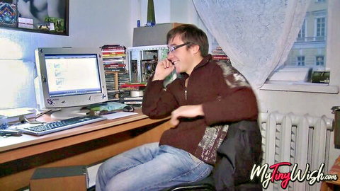 Smiling man in glasses talks on phone at cluttered desk with computer in dorm room