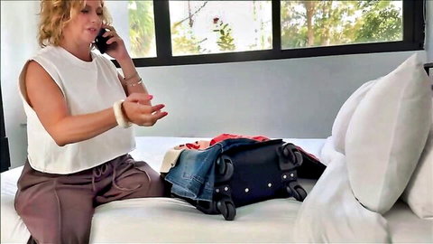 Blonde MILF in tight white top and pants sits on bed talking on phone with open suitcase