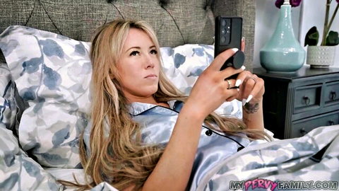Blonde stepmom in silky pajamas holding phone on bed, masturbation tease
