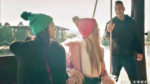 Two girls in colorful beanies sit on outdoor swing with smiling guy behind in casual threesome tease