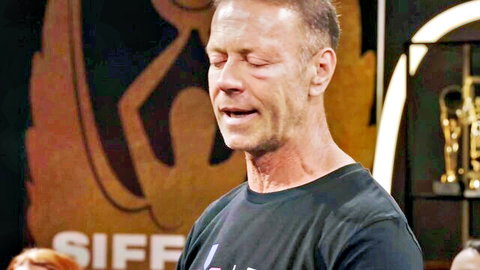 Bald middle-aged man passionately speaking on stage with trophies and emblem backdrop