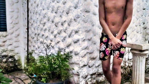 Shy shirtless young man in floral swim trunks covering crotch outdoors
