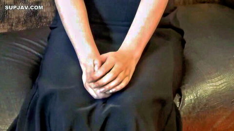 Sexy Japanese woman in tight black dress sits on couch with hands clasped over lap