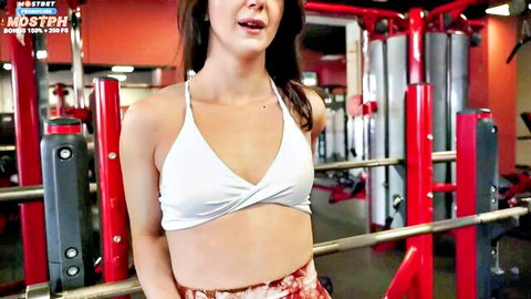 Brunette gym babe in skimpy white bikini top flaunts cleavage and toned abs near red pull-up bars