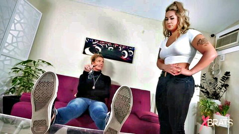 Blonde femdom mistress stands dominant over seated woman with Converse soles up in lesbian foot worship tease