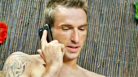 Muscular tattooed blonde hunk shirtless talking on phone in tropical hut