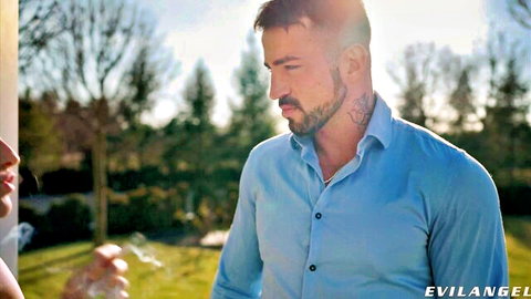 Tattooed hunk in blue shirt stands with smoking woman's lips near him outdoors