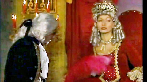 Busty blonde queen in red gown and crown flirts with kneeling powdered wig man holding pink fan
