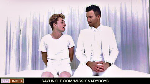 Blonde twink in white shorts and suited man staring intensely on bed in Missionary Boys scene