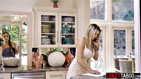 Busty blonde MILF with big tits talks on phone in kitchen while brunette babe watches in mirror