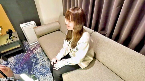 Cute Japanese teen in white sweater smiling teasingly on hotel couch