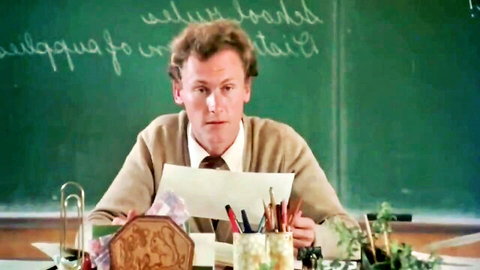 Mature teacher in cardigan at desk, classroom roleplay scene