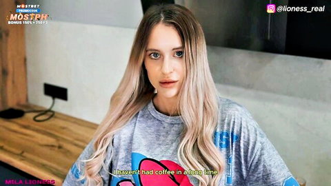 Young blonde babe in oversized gray t-shirt posing casually on desk craving coffee.