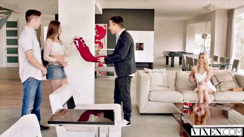 Suited agent hands red scarf to sexy brunette in crop top and mini skirt while blonde model lounges on couch