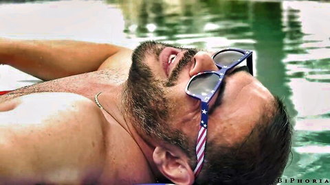 Bearded man lounging shirtless by pool in patriotic shades, BiPhoria video with Aften Opal & Rose Lynn