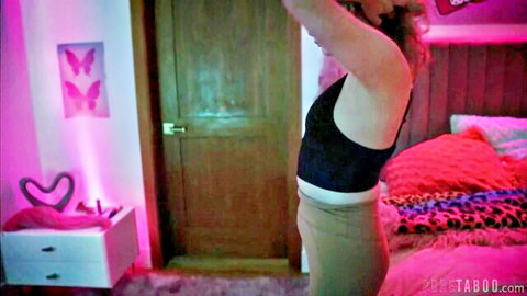 Blonde girl stretching arms up in black tank top and low pants exposing midriff in pink neon bedroom
